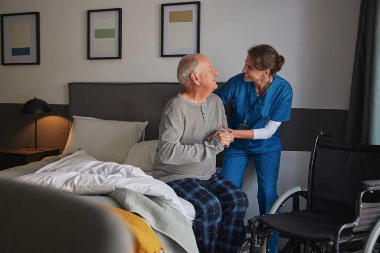 Caregiver respectfully assists an older resident in a bright, tidy assisted living common area.